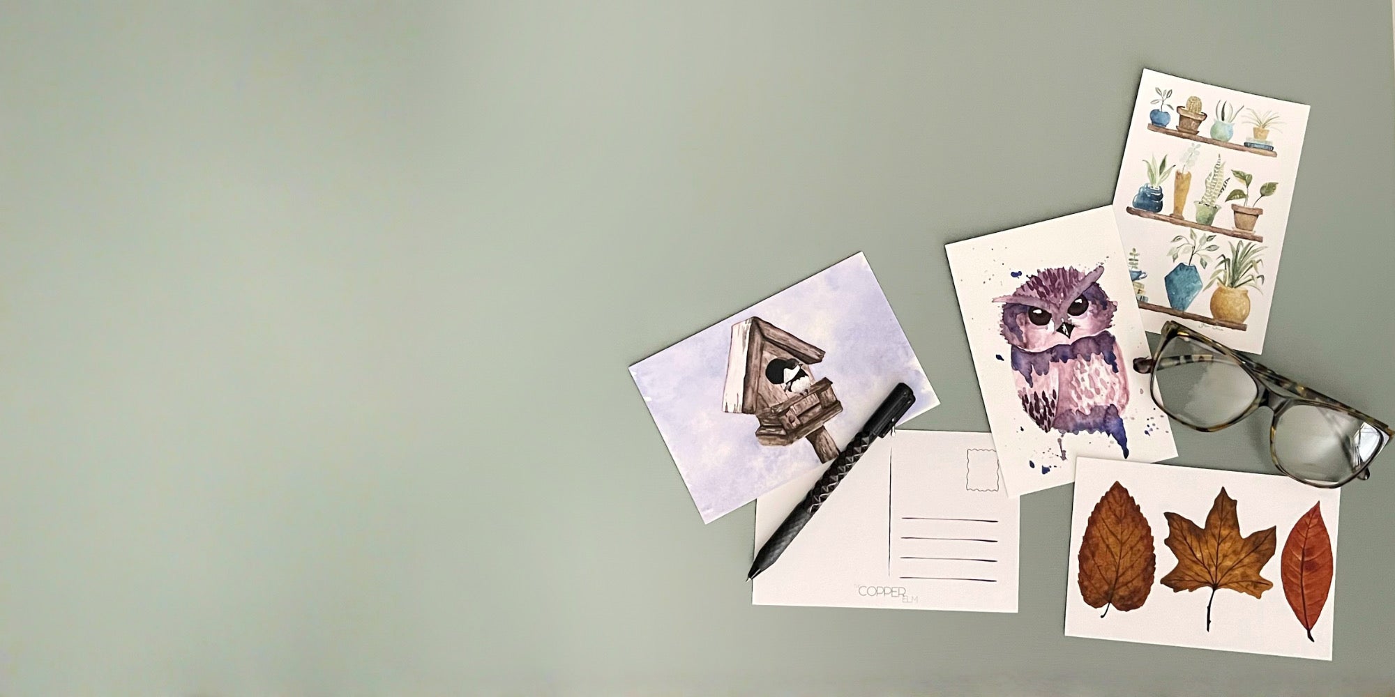 Five postcards with watercolor artwork sitting on desk with pen and reading glasses