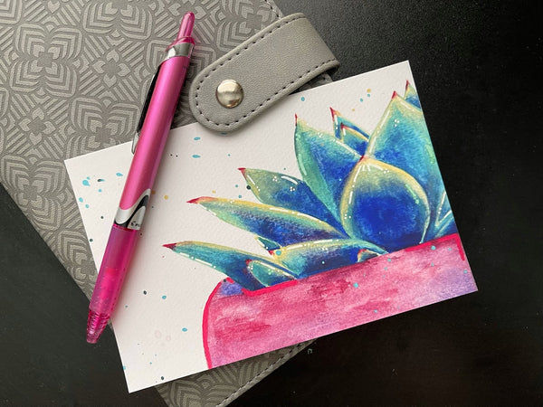 Postcard print of watercolor painting of a vibrantly colored succulent sitting on a gray notebook with a pink pen