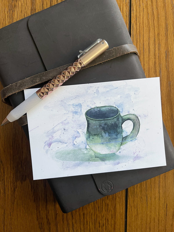 Watercolor painting postcard of a green pottery mug, photographed on a leather notebook with pen.