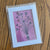 Matted artwork of a floral bouquet on a pink background in a protective clear sleeve, displayed on a wooden surface.