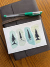 Water color postcard of pine trees on green background sitting on leather notebook. 