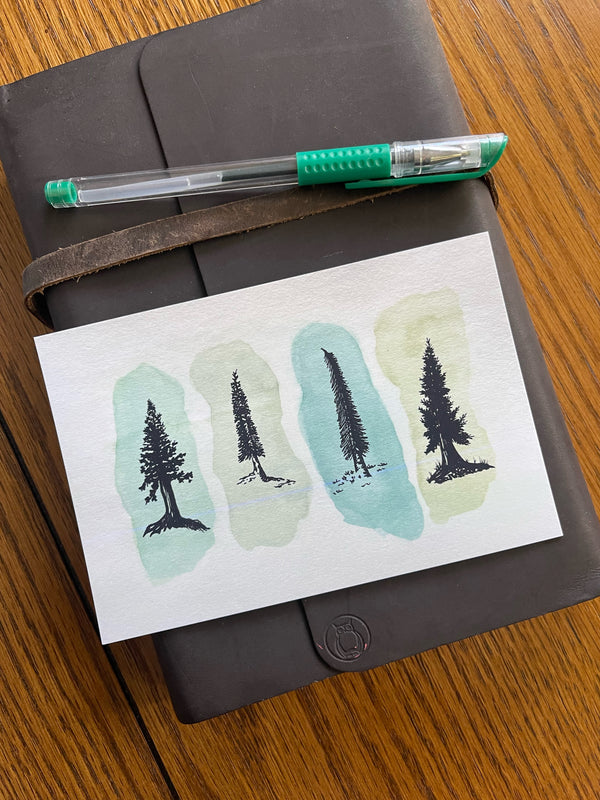 Water color postcard of pine trees on green background sitting on leather notebook. 